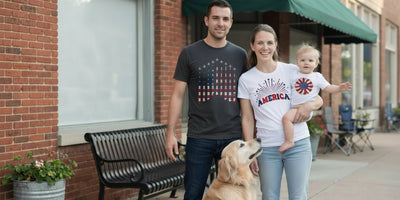 A young family on a sunlit sidewalk with their dog, wearing Proud & Free airplane smoke and America fireworks tees