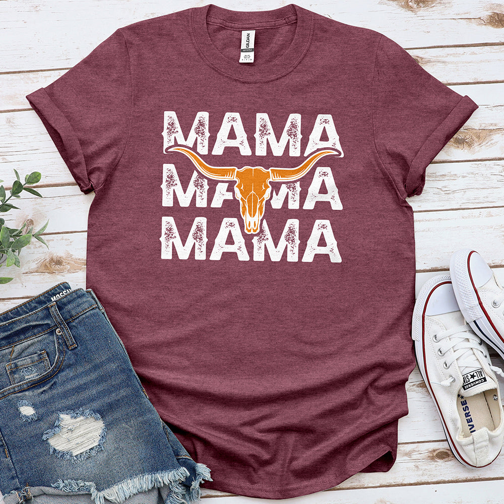 Heather maroon t-shirt with stacked 'MAMA' text and a longhorn skull graphic, laid out with denim shorts and white sneakers on a wooden background.