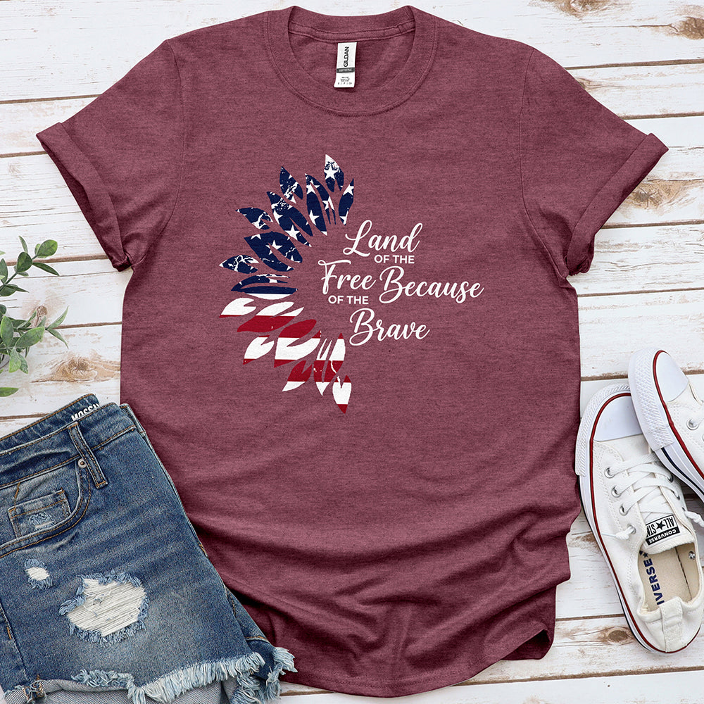 Heather maroon patriotic graphic tee with 'Land of the Free Because of the Brave' text and USA flag feather design, styled with denim shorts and white sneakers on wood background.