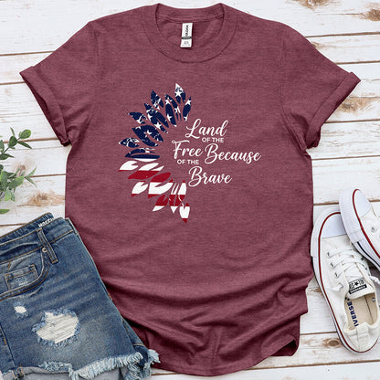 Heather maroon patriotic graphic tee with 'Land of the Free Because of the Brave' text and USA flag feather design, styled with denim shorts and white sneakers on wood background.