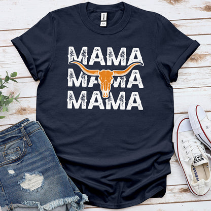 Navy blue t-shirt with distressed 'MAMA' text and longhorn skull graphic, styled with denim shorts and white sneakers on wooden background.