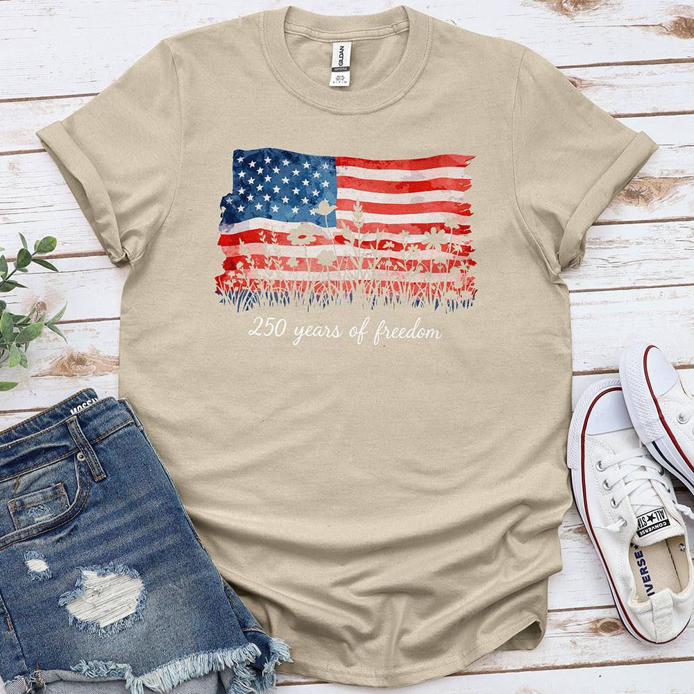 Beige t-shirt with American flag and '250 years of freedom' graphic, laid flat with denim shorts and white sneakers.