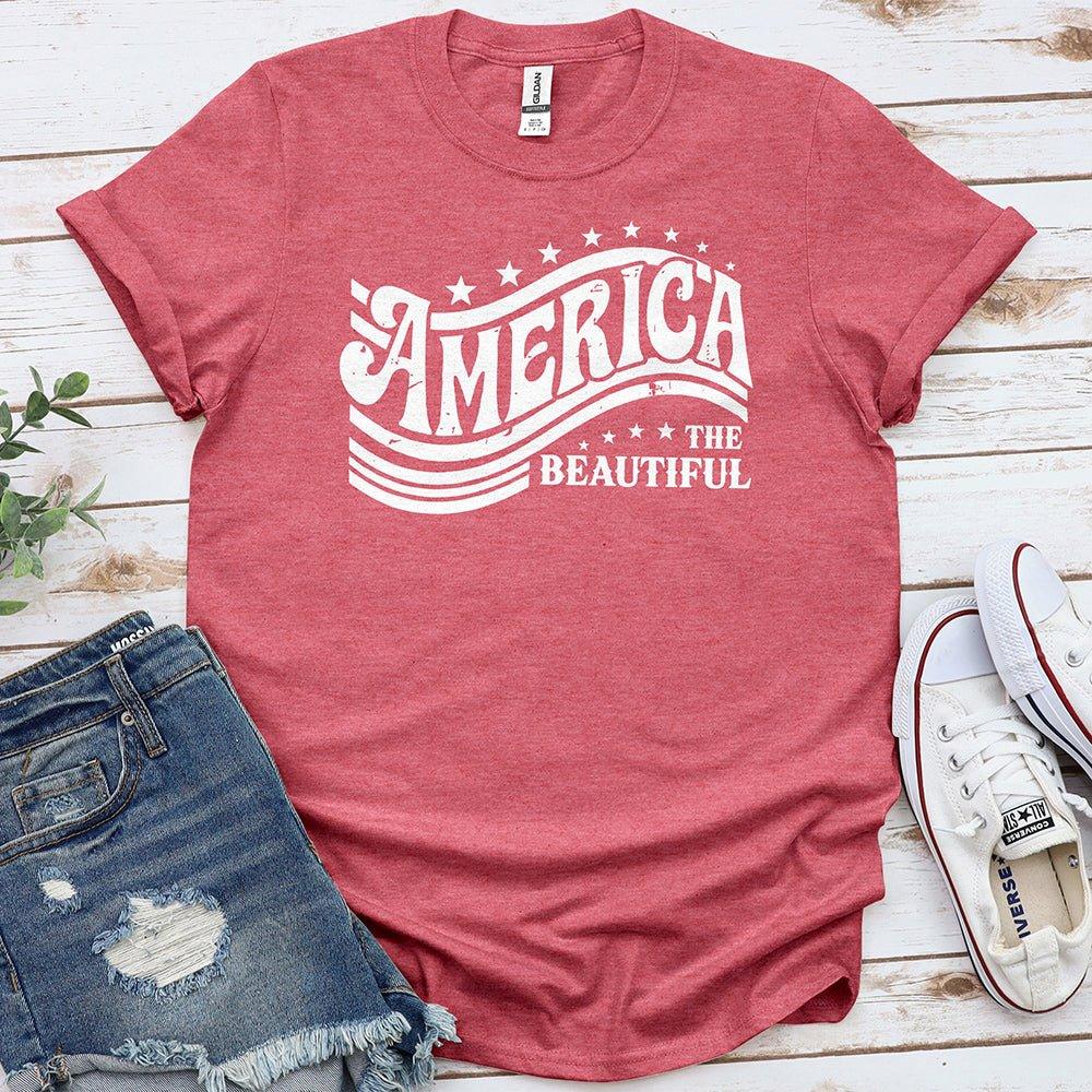 Red 'America the Beautiful' graphic tee with denim shorts and white sneakers on wood.