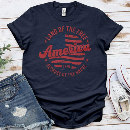 Navy blue "Land of the Free America Because of the Brave" 1776 patriotic t-shirt flat lay with denim shorts and white Converse shoes on wood background.