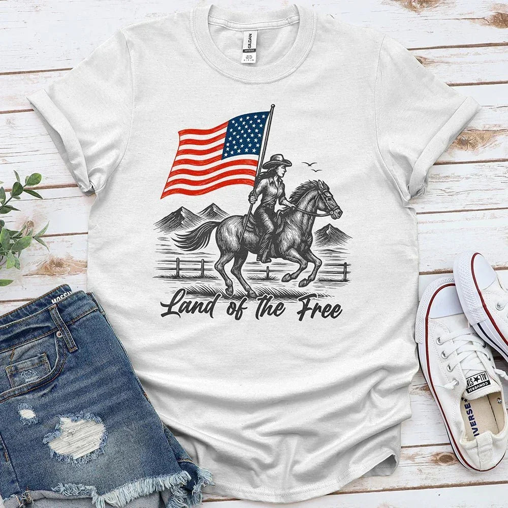 White t-shirt with 'Land of the Free' cowboy, horse, and American flag graphic, laid with denim shorts and sneakers.