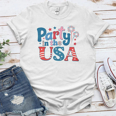 Party in the USA graphic tee with patriotic red, white, and blue design, styled with denim shorts and sneakers on wood background.