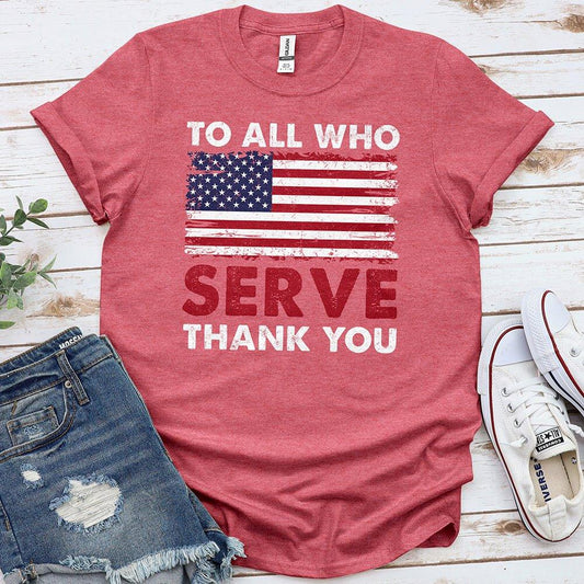 Red Proud & Free T-shirt with American flag and 'To All Who Serve Thank You' message, styled with denim shorts and white sneakers on wood background.